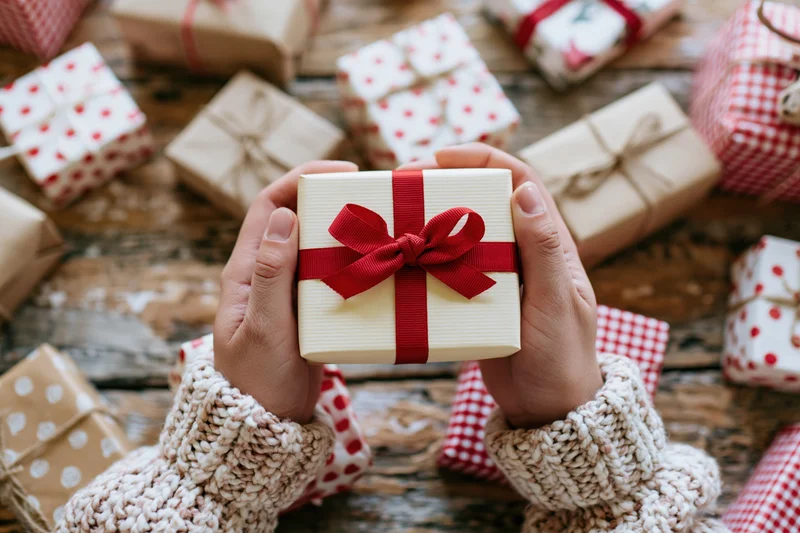 Hands holding a cream gift box with red ribbon over a wooden table surrounded by wrapped presents
