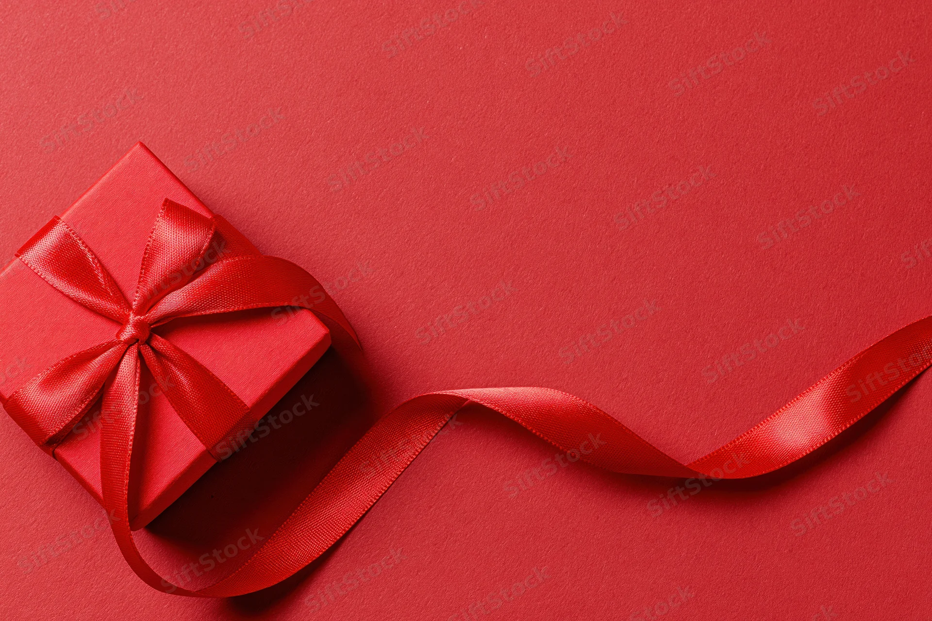 Red gift box wrapped in satin ribbon tied into a bow on a red surface with trailing ribbon and copyspace
