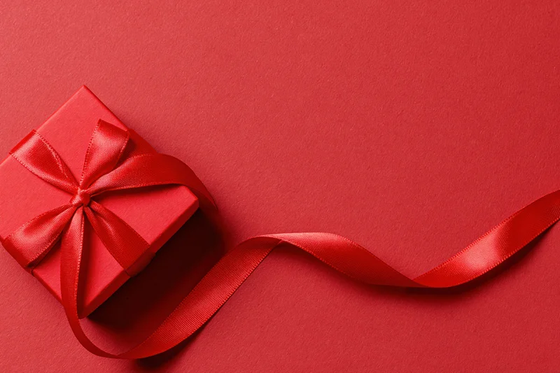 Red gift box wrapped in satin ribbon tied into a bow on a red surface with trailing ribbon and copyspace
