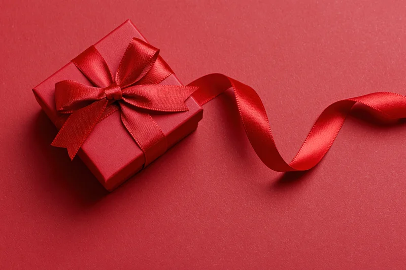 Red gift box wrapped in satin ribbon tied into a bow on a red surface with trailing ribbon and copyspace