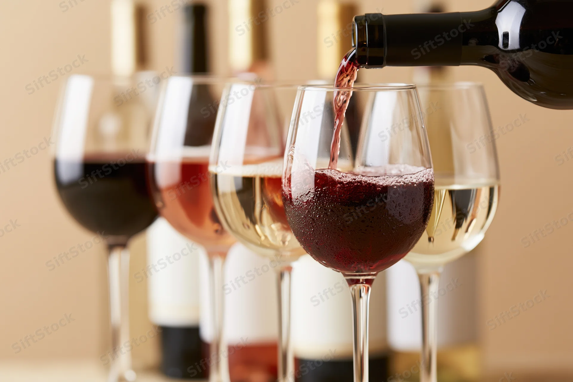 Red wine being poured from a dark bottle into the foremost of a row of wine glasses with other glasses and bottles in the background