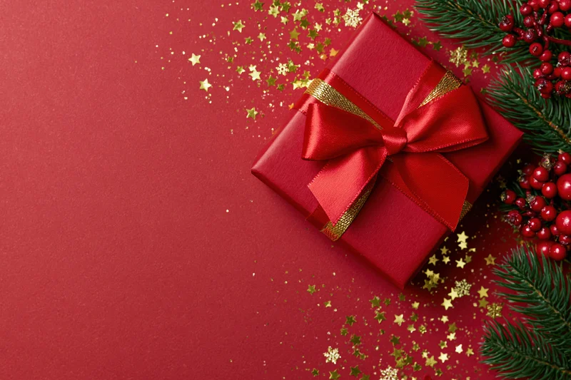 Red wrapped gift box with a large satin bow and pine branches on a red surface