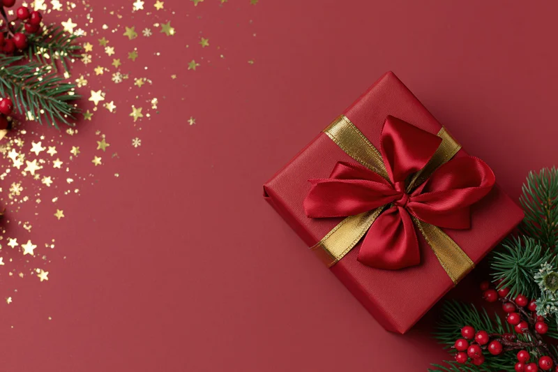 Red wrapped gift box with a large red satin bow and gold ribbon on a red surface, surrounded by pine branches, red berries, and gold star confetti, with copyspace on the left