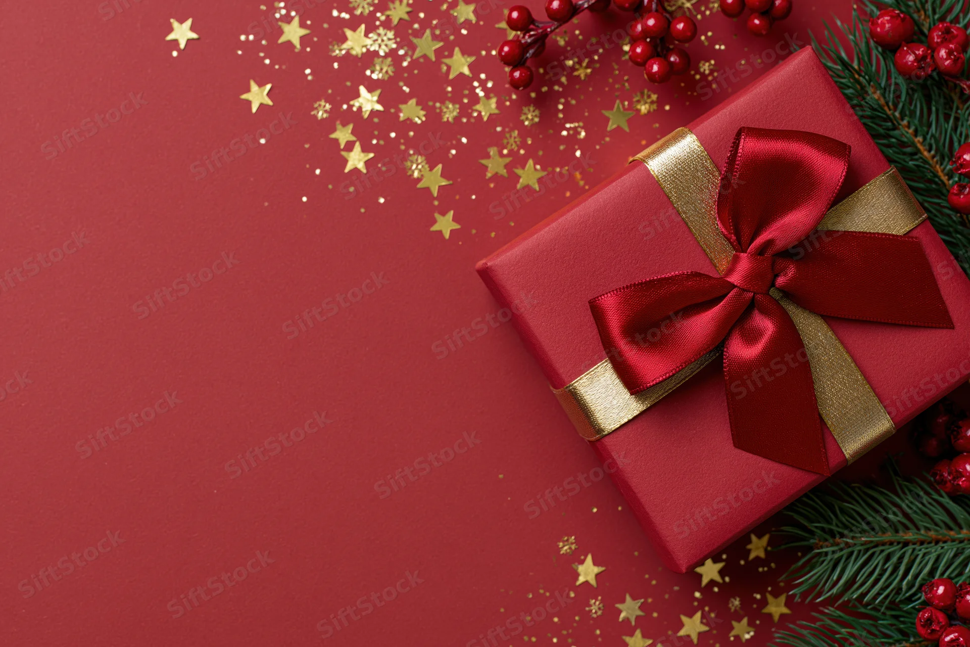Red gift box with a large red satin bow and gold ribbon on a red surface surrounded by pine branches, red berries, and gold star confetti