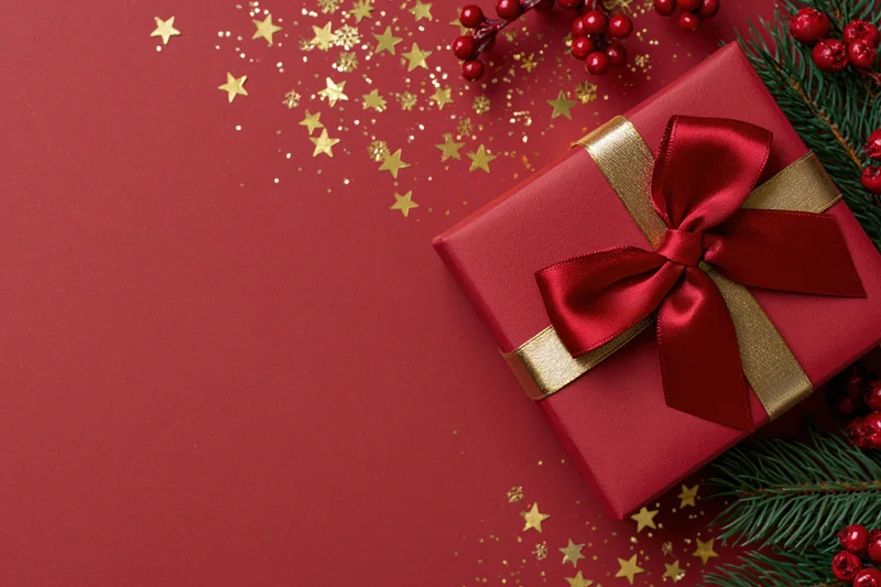 Red gift box with a large red satin bow and gold ribbon on a red surface surrounded by pine branches, red berries, and gold star confetti