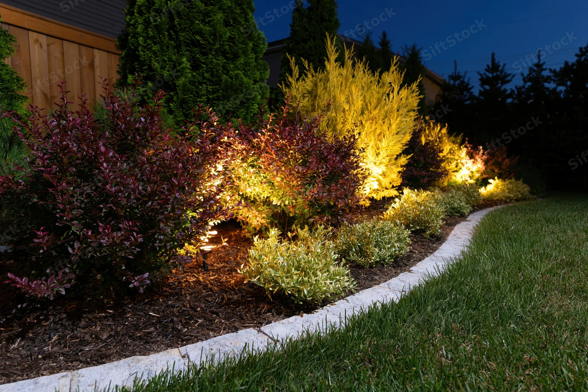 Illuminated garden bed with shrubs and mulch along a curved stone border at dusk