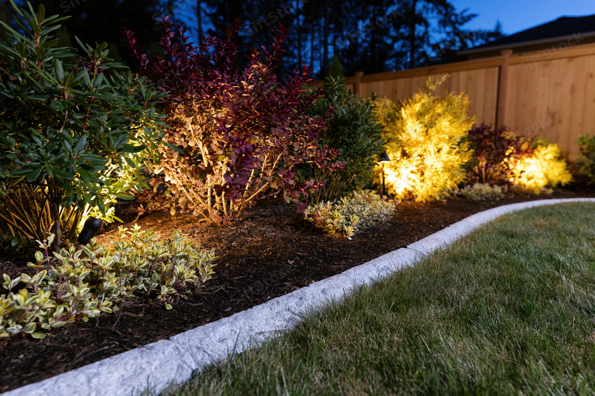 Illuminated garden bed with shrubs and mulch along a grassy lawn at dusk