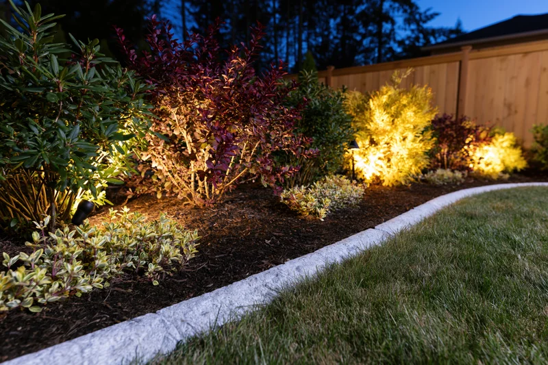 Illuminated garden bed with shrubs and mulch along a grassy lawn at dusk
