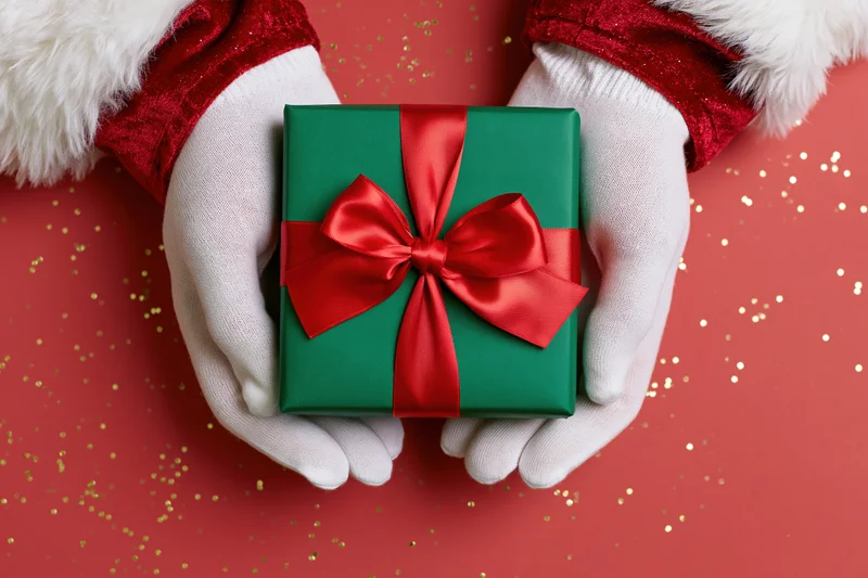 Santa's gloved hands holding a green gift box with red ribbon on red glitter background