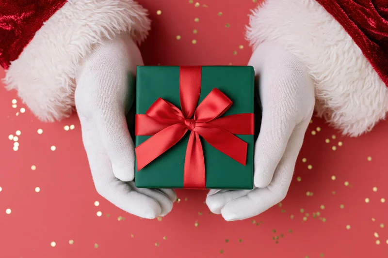 Santa's gloved hands holding a green gift box with red ribbon on red background with gold confetti