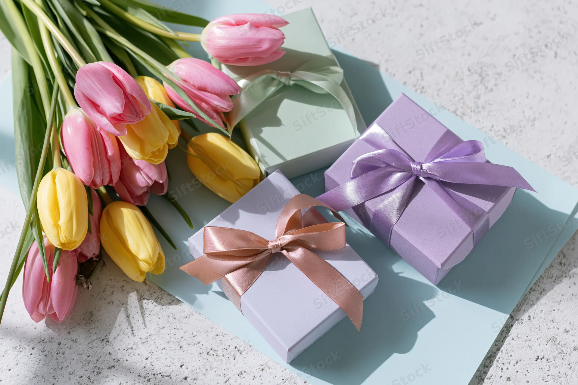 Pastel-wrapped gift boxes with satin ribbon bows and a bouquet of pink and yellow tulips on a light blue surface