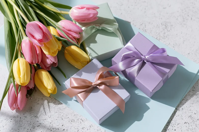 Pastel-wrapped gift boxes with satin ribbon bows and a bouquet of pink and yellow tulips on a light blue surface