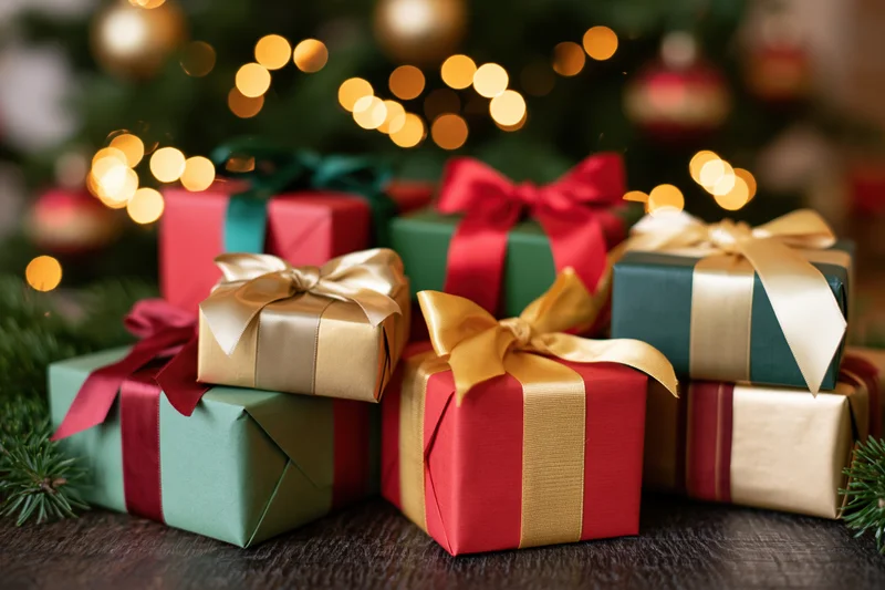 Wrapped christmas gifts in red green and gold arranged on a wooden table with blurred tree lights in background