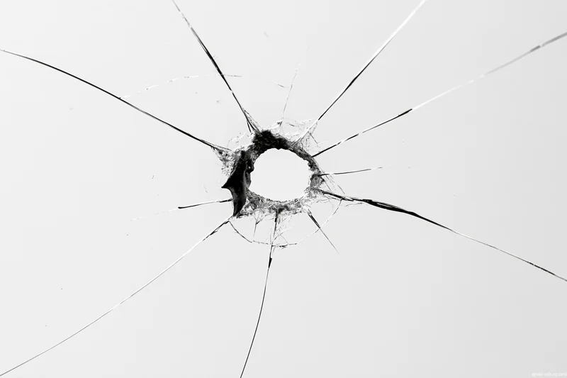 Shattered glass pane with central impact hole and radiating cracks against white background