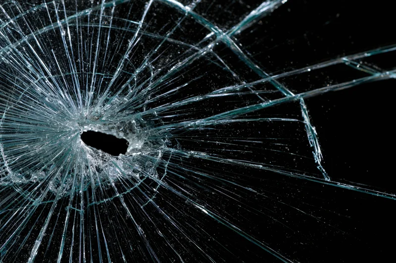 Shattered glass pane with central impact hole and radiating cracks against dark background