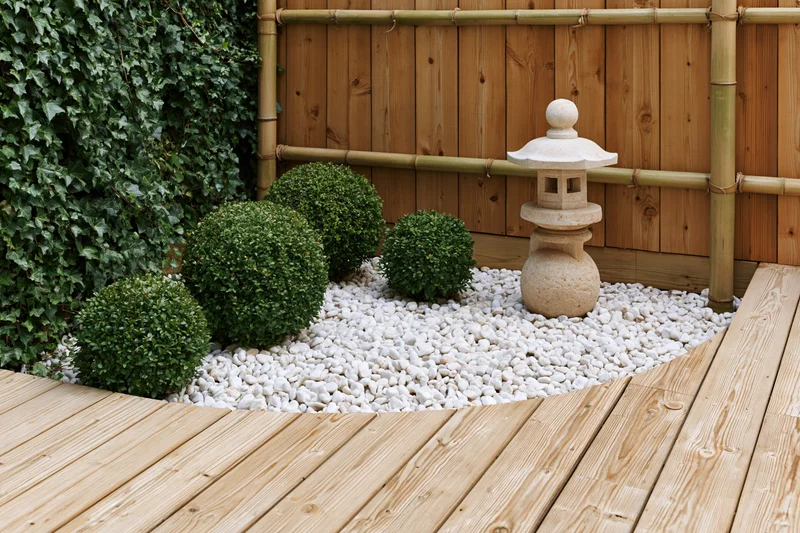 Japanese-style garden corner with stone lantern and trimmed boxwood shrubs on white pebbles beside wooden deck