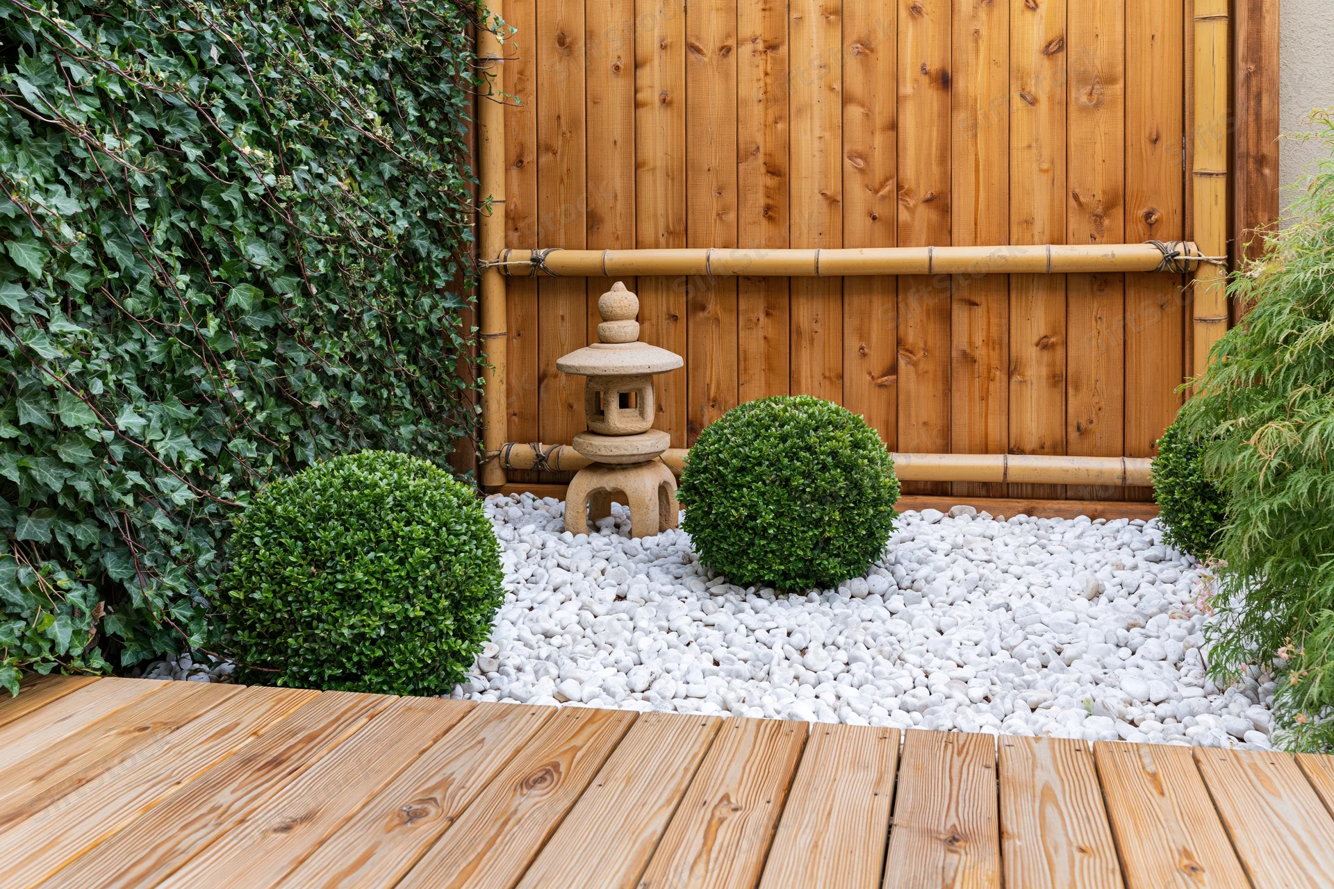 Japanese-style courtyard garden with stone lantern and trimmed boxwood shrubs on white pebbles beside wooden deck and bamboo fence