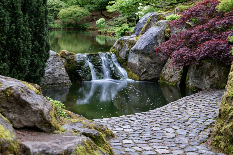 Small cascading waterfall flowing over mossy granite boulders into a pond beside a cobblestone path in a Japanese garden