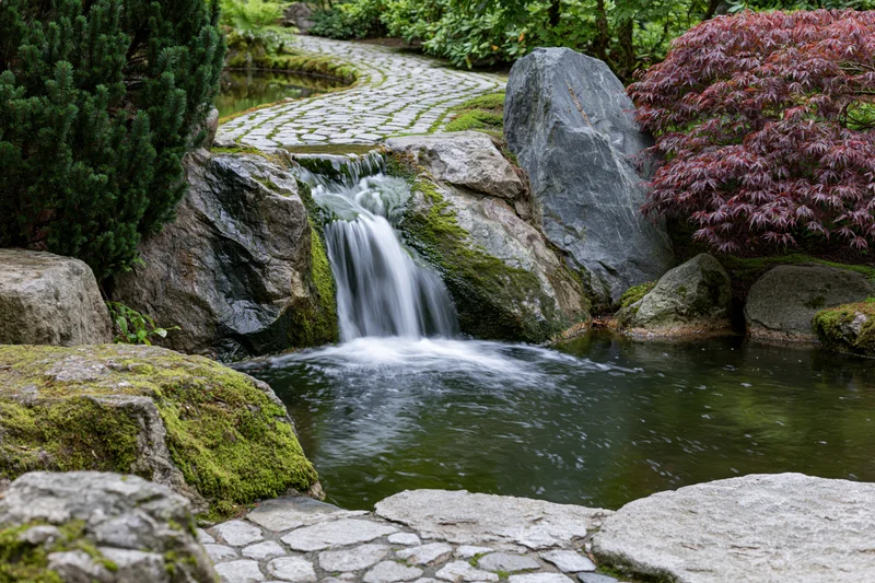 Small cascading waterfall flowing over mossy granite boulders in a Japanese garden with a stone path and red maple tree