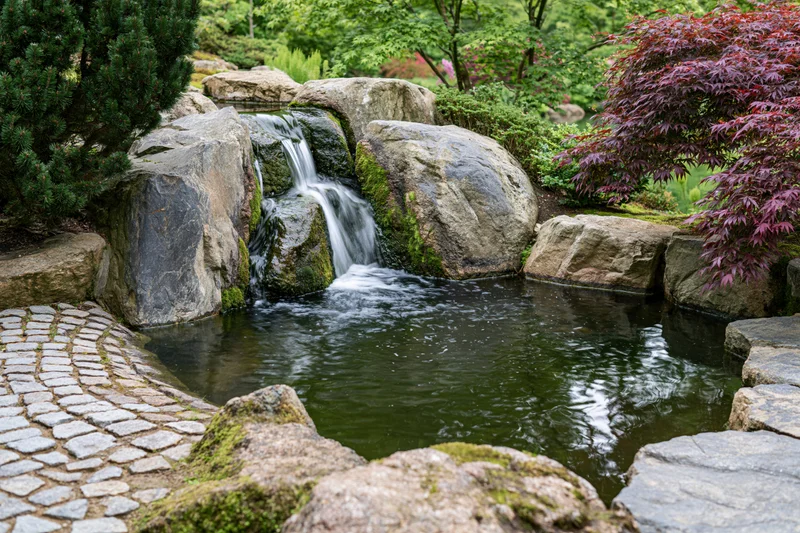 Small cascading waterfall flowing over mossy granite boulders into a pond in a Japanese garden with cobblestone path and surrounding foliage