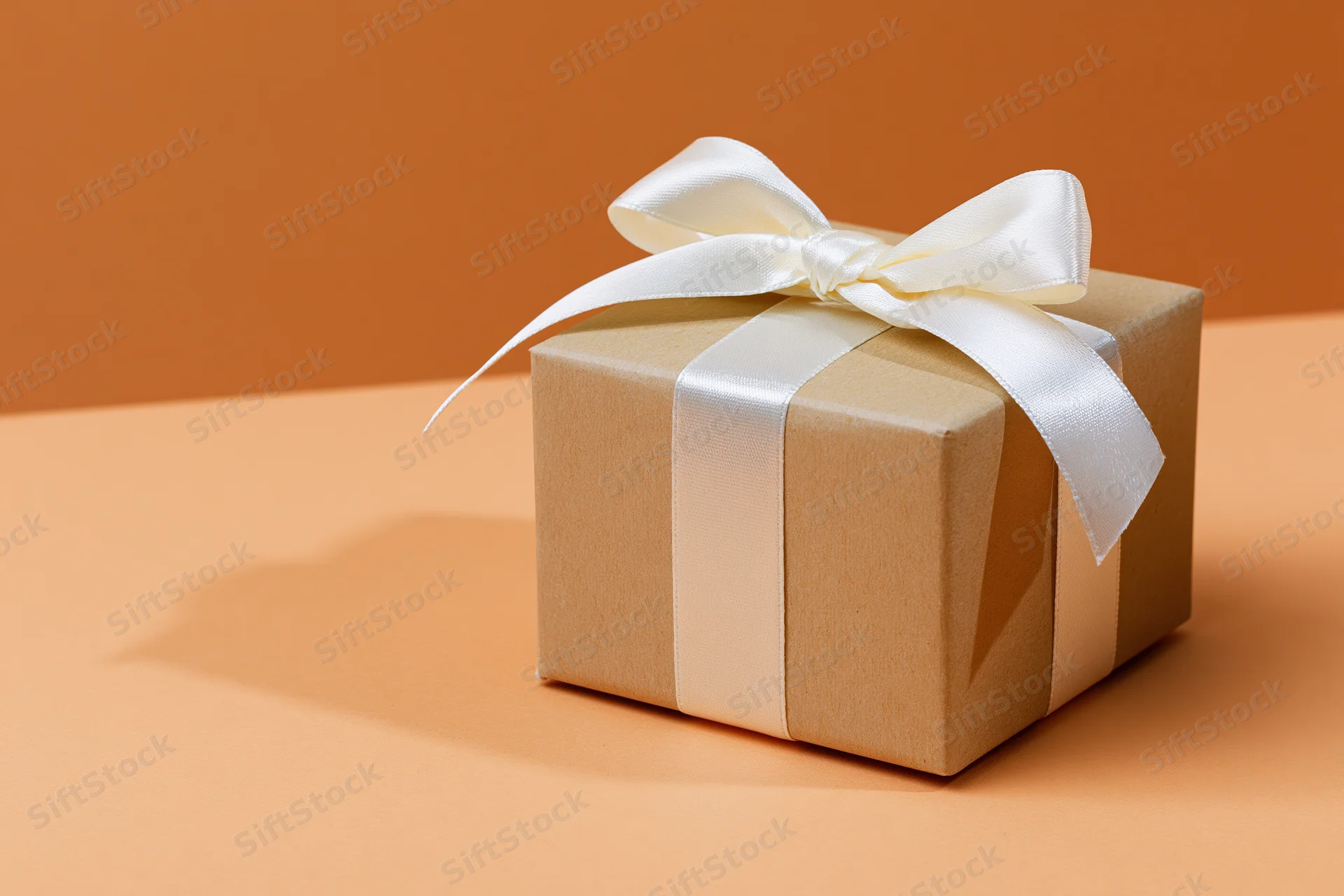 Small kraft cardboard gift box tied with white satin ribbon on orange surface