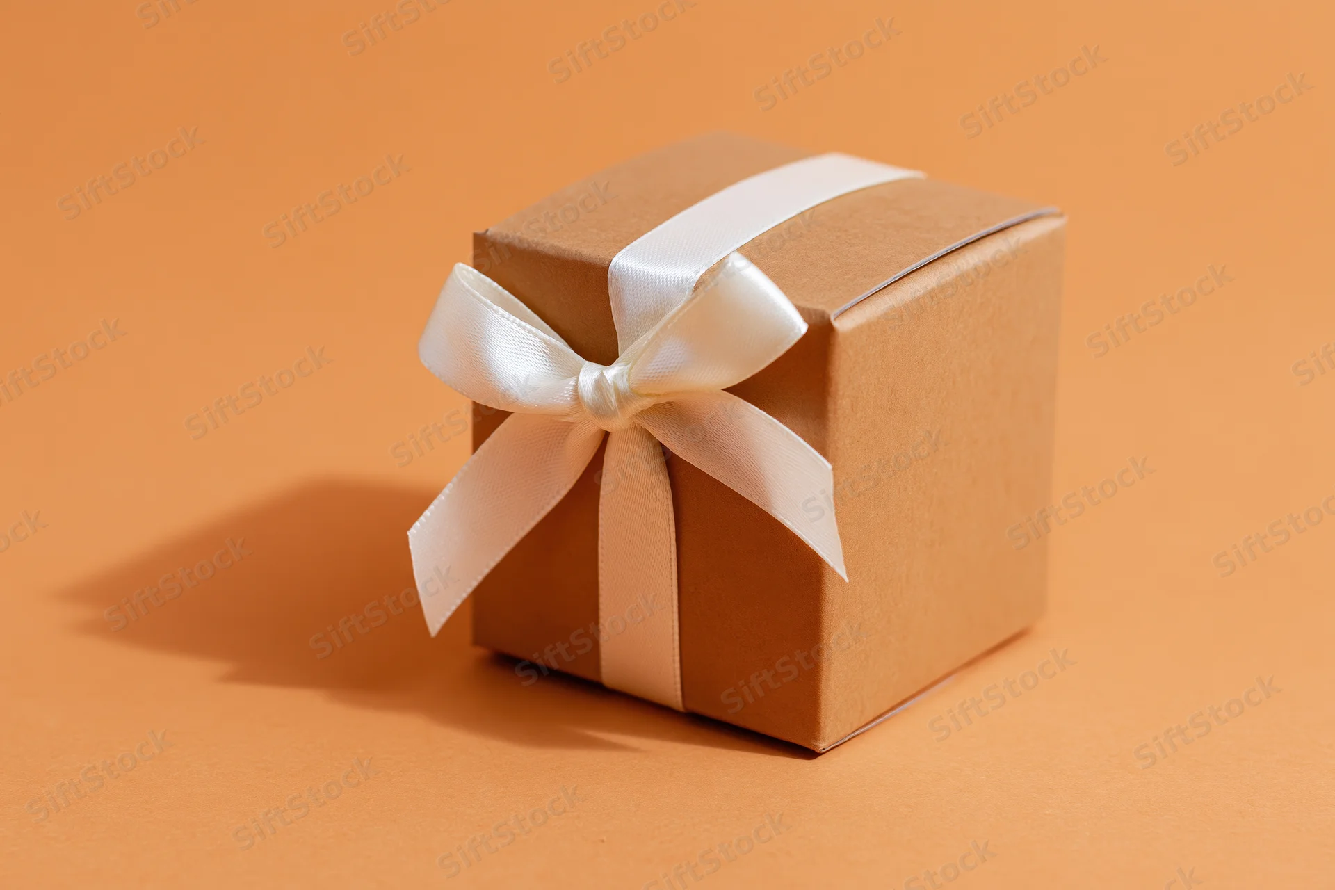 Small kraft cardboard gift box tied with white satin ribbon on orange background