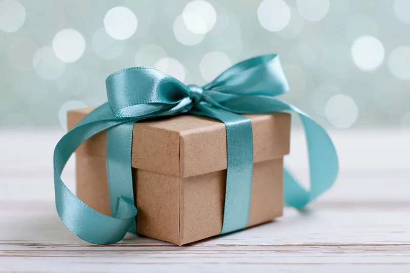 Small kraft paper gift box tied with a satin teal ribbon on a wooden surface with bokeh lights in the background
