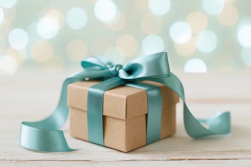 Small kraft paper gift box tied with a satin teal ribbon on a wooden surface with blurred festive lights in the background