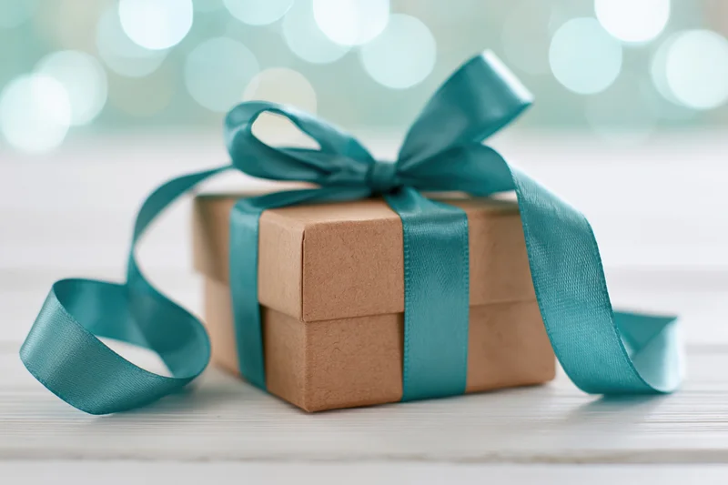 Small kraft paper gift box tied with a satin teal ribbon on a white wooden surface with blurred bokeh lights in the background