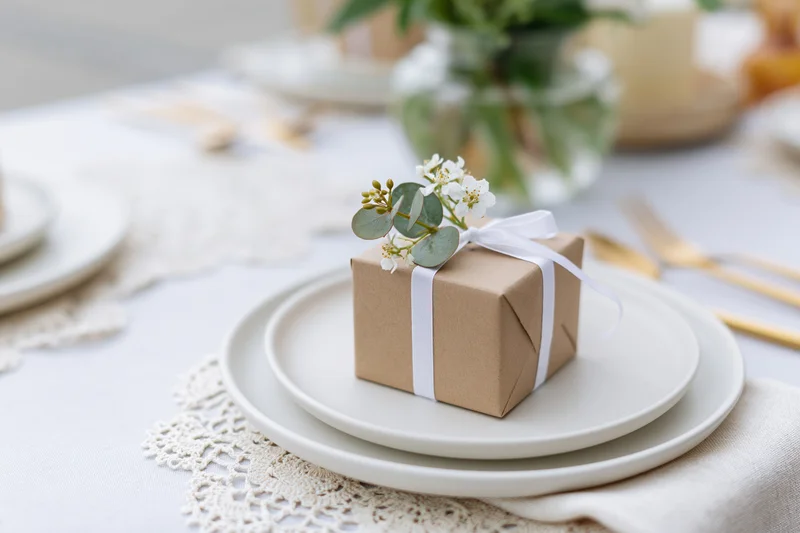 Small kraft paper gift box tied with white ribbon and eucalyptus flower on stacked white plates at a table setting
