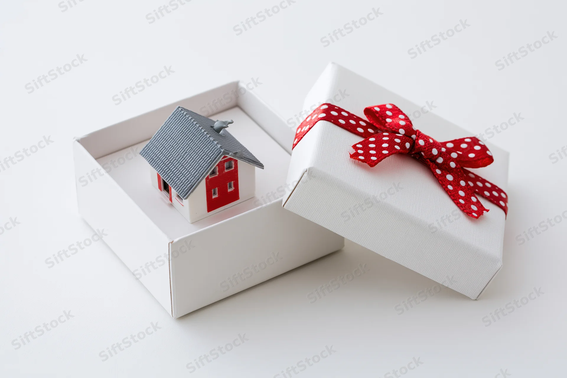 Miniature house model inside open gift box with red polka dot ribbon on white background