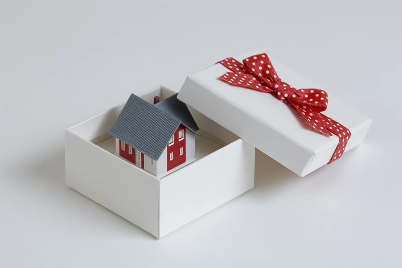 Miniature house model inside open gift box with red polka dot ribbon on white background