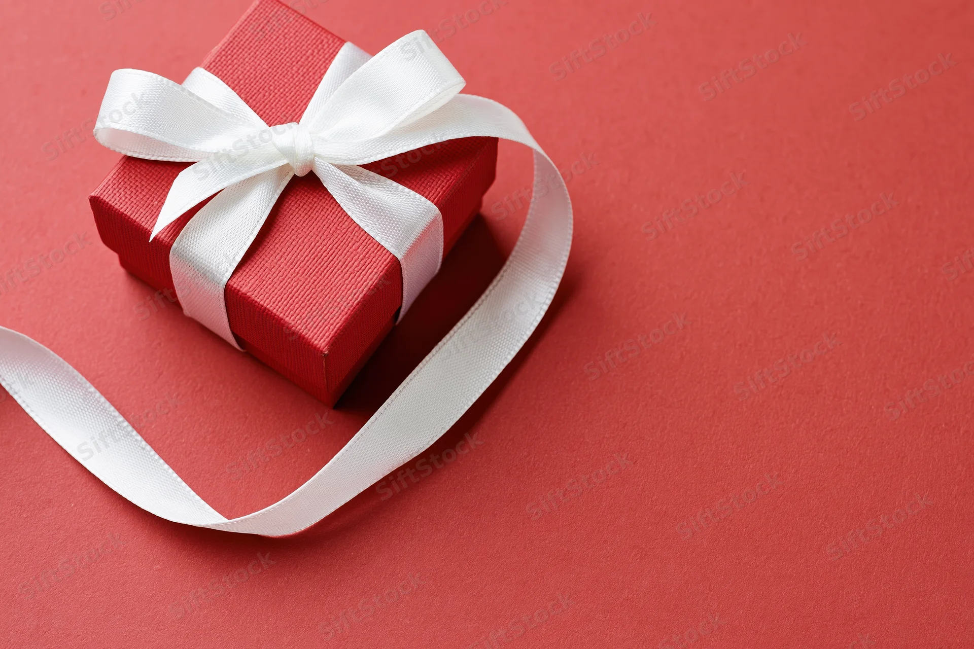 Small red textured gift box with white satin ribbon bow on red surface