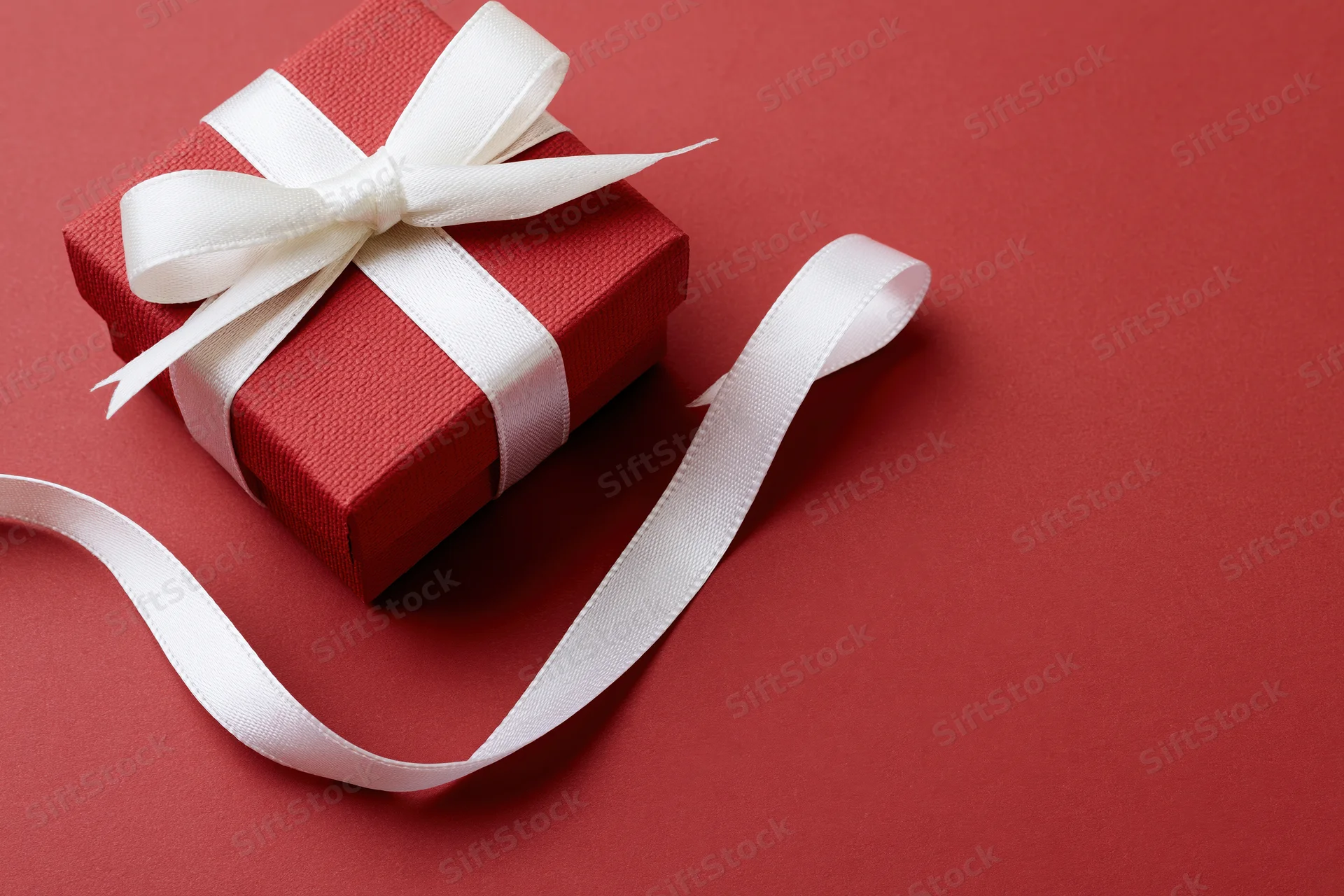 Small red textured gift box with white satin ribbon bow on red surface