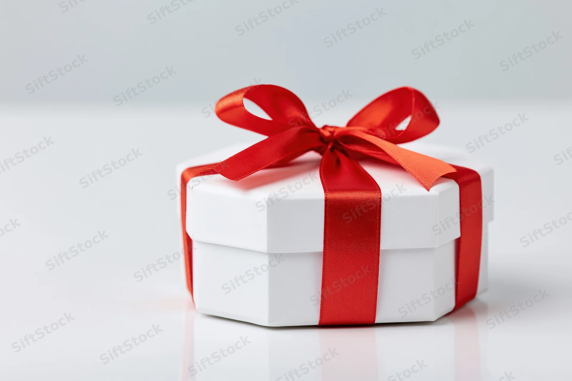 White octagonal gift box tied with red satin ribbon on reflective white surface