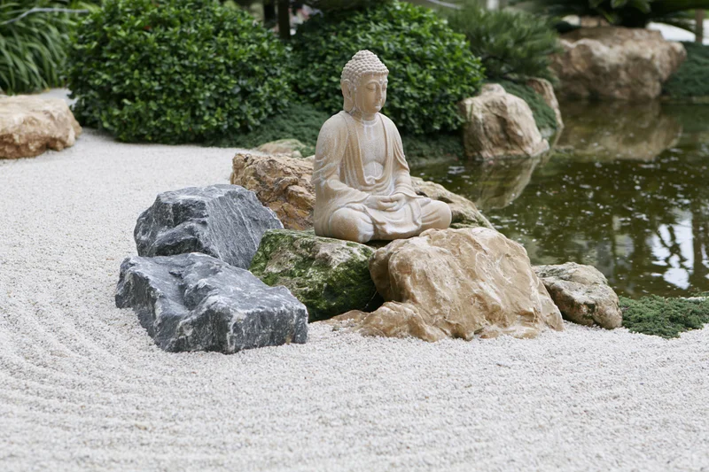 Stone buddha statue seated on rocks in a raked gravel zen garden with water and greenery
