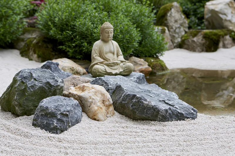 Seated stone buddha statue in a zen garden with raked white gravel, rocks, and green shrubs near a small water feature