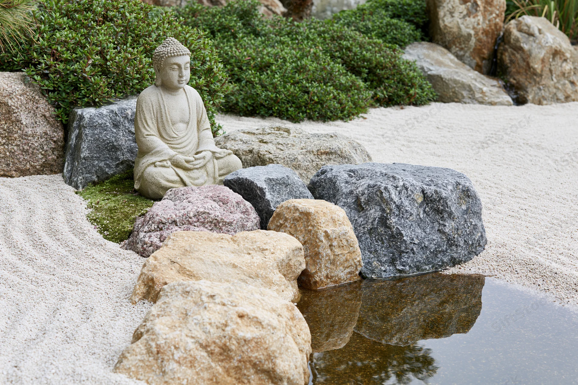 Seated stone buddha statue in a zen garden with raked gravel, rocks, and a small water feature