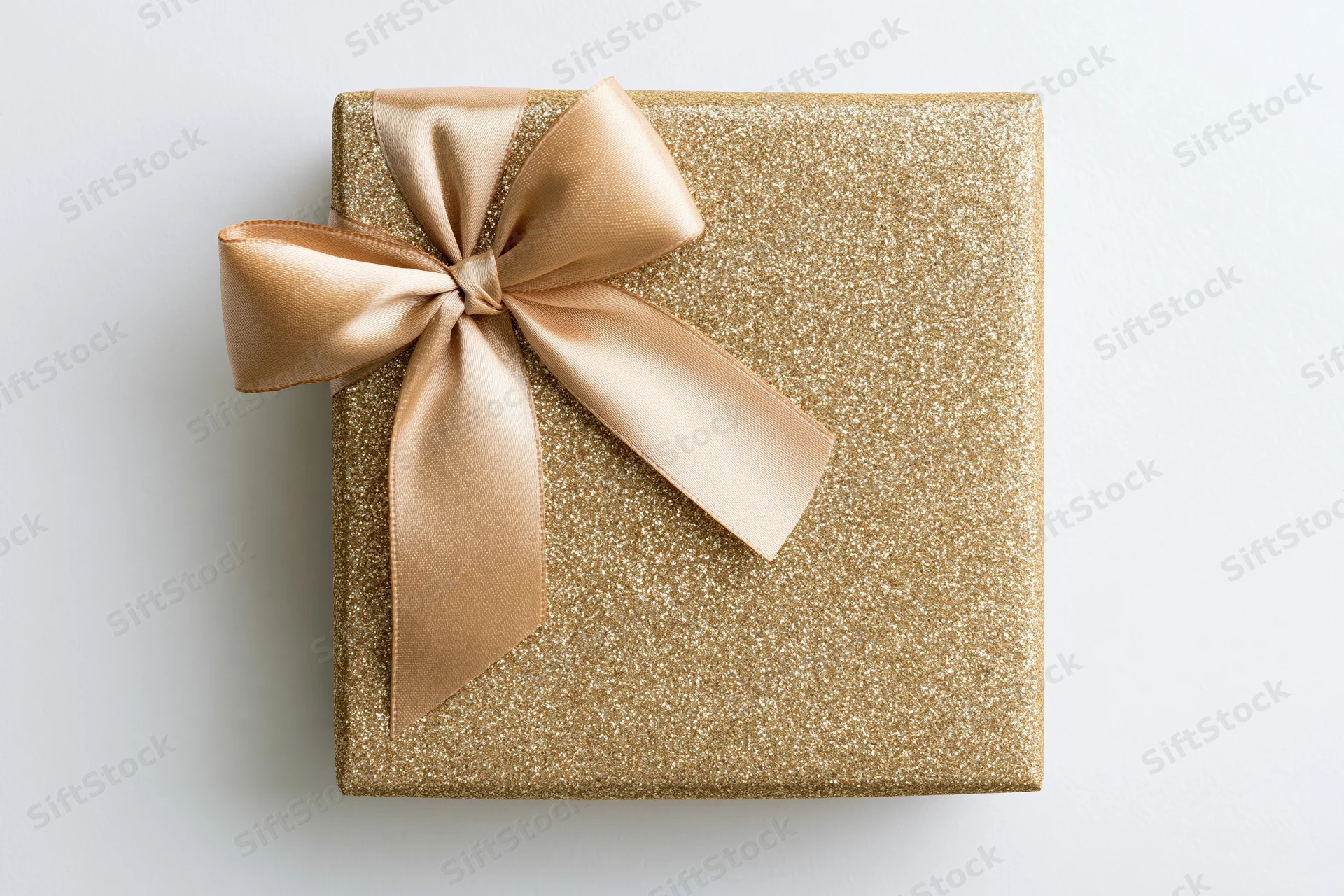 Square gift box wrapped in gold glitter paper with a satin ribbon bow on white surface