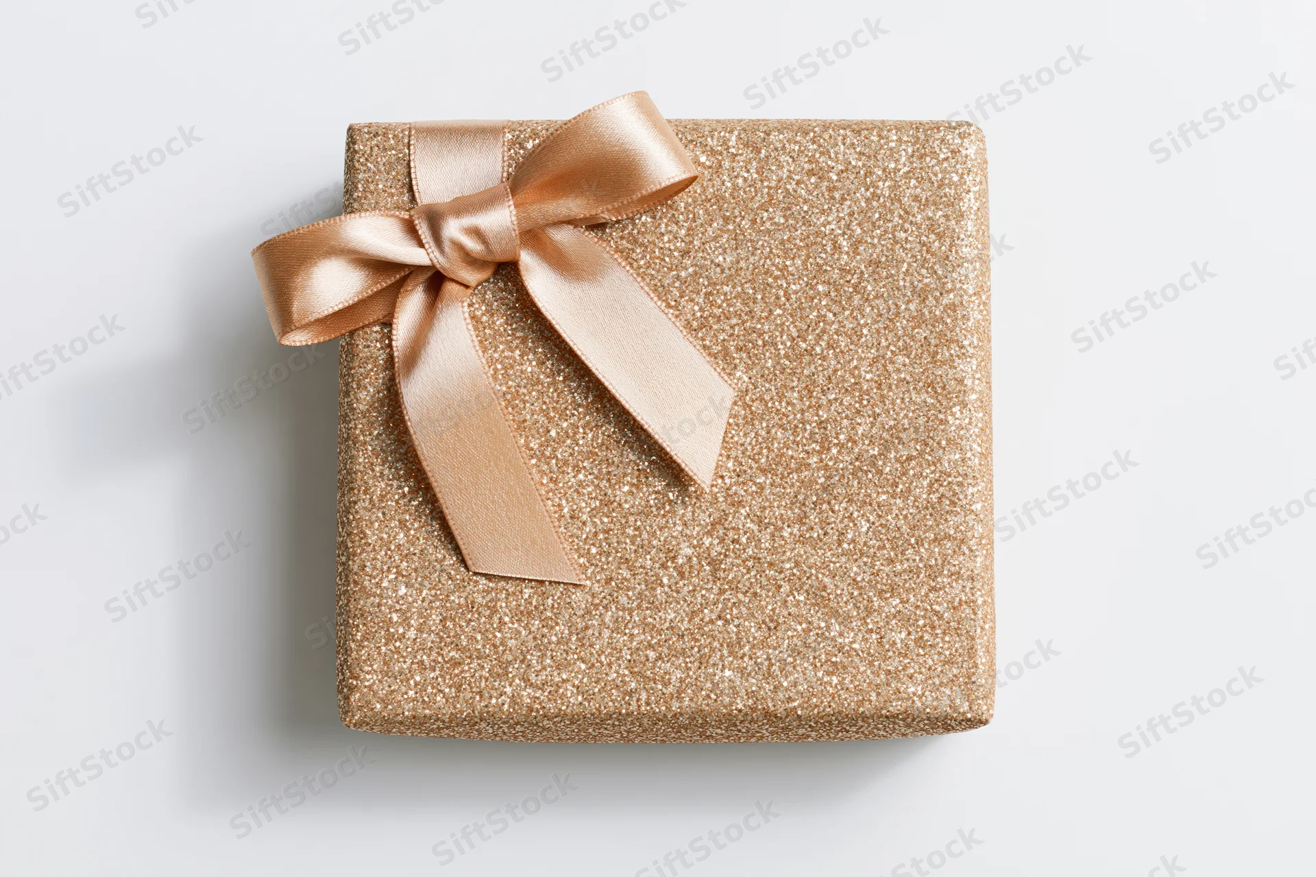 Square gift box wrapped in gold glitter paper with a satin ribbon bow on white background
