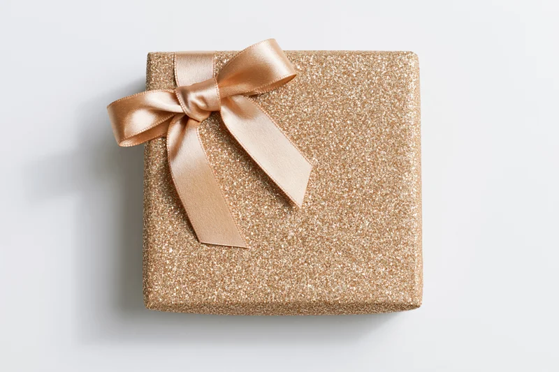 Square gift box wrapped in gold glitter paper with a satin ribbon bow on white background