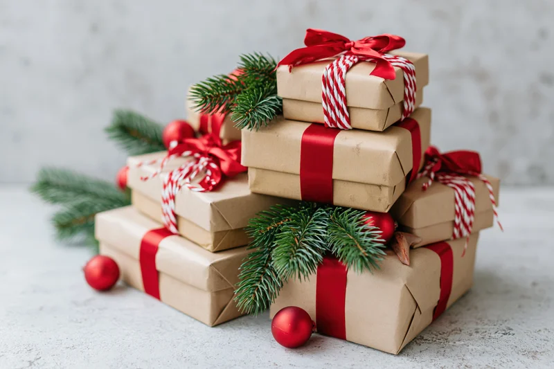 Stack of christmas gifts wrapped in kraft paper with red ribbons and pine branches on a light surface