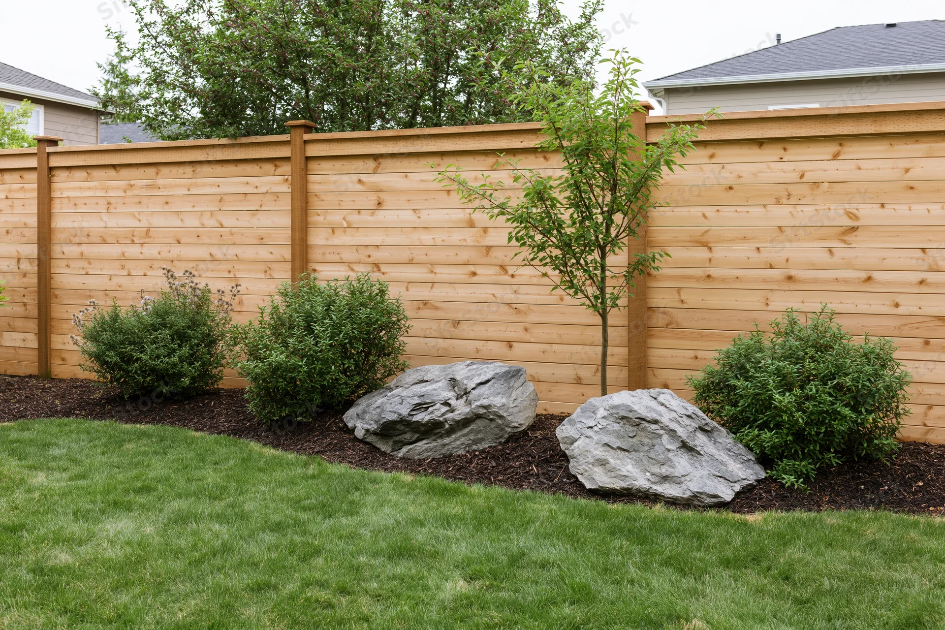 Suburban backyard garden bed with ornamental shrubs, small tree, rocks and mulch along wooden fence