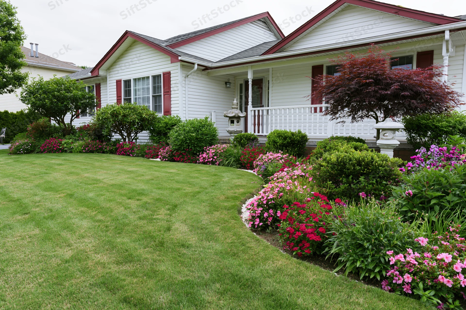 White suburban house with red trim and manicured front yard featuring green lawn, flower beds, and shrubs