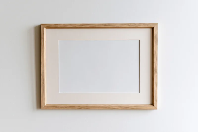 Light oak wooden picture frame with blank white interior hanging on a white wall