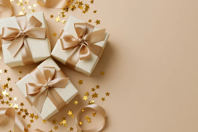 Three cream gift boxes with gold satin ribbons and star confetti on beige surface