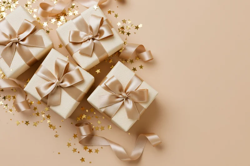 Four cream gift boxes with gold satin ribbons and star confetti on beige surface