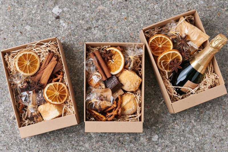 Three open kraft gift boxes filled with dried oranges, cinnamon, star anise, chocolate, biscuits and champagne on a gray concrete surface