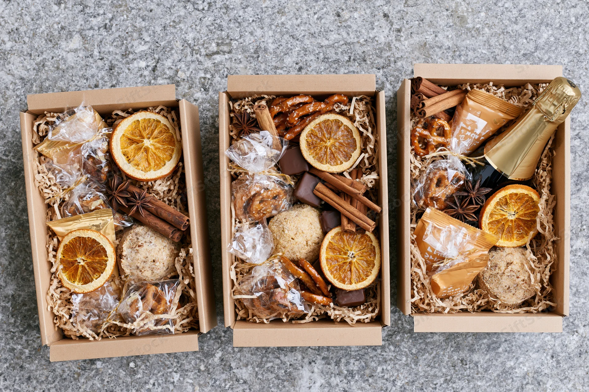 Three open kraft gift boxes filled with assorted holiday treats and dried fruit arranged on a stone surface