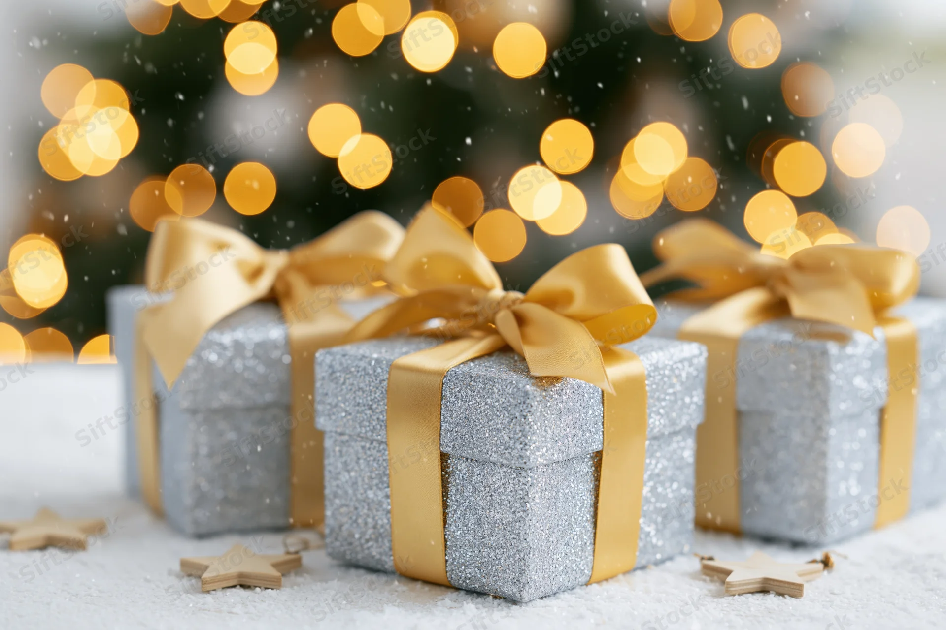 Three silver glitter gift boxes with gold satin ribbon bows arranged on snowy surface near blurred holiday lights